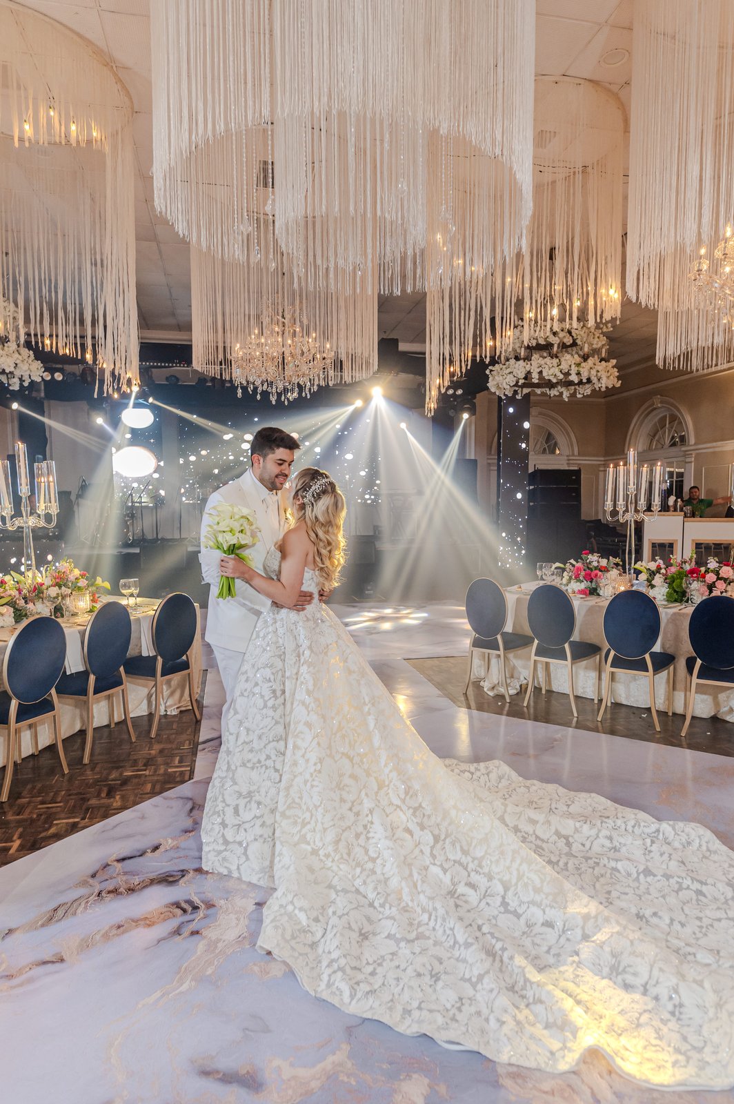 Novia con vestido de cola larga abrazada por el novio en recepción con chandeliers de hilos