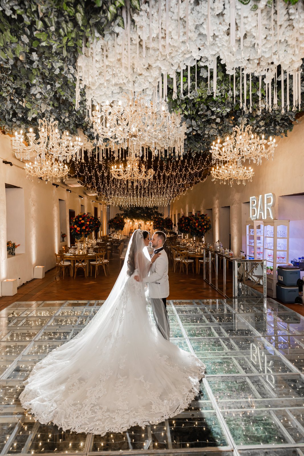Primer baile sobre piso de vidrio iluminado bajo techo de flores y chandeliers en boda de Cartagena